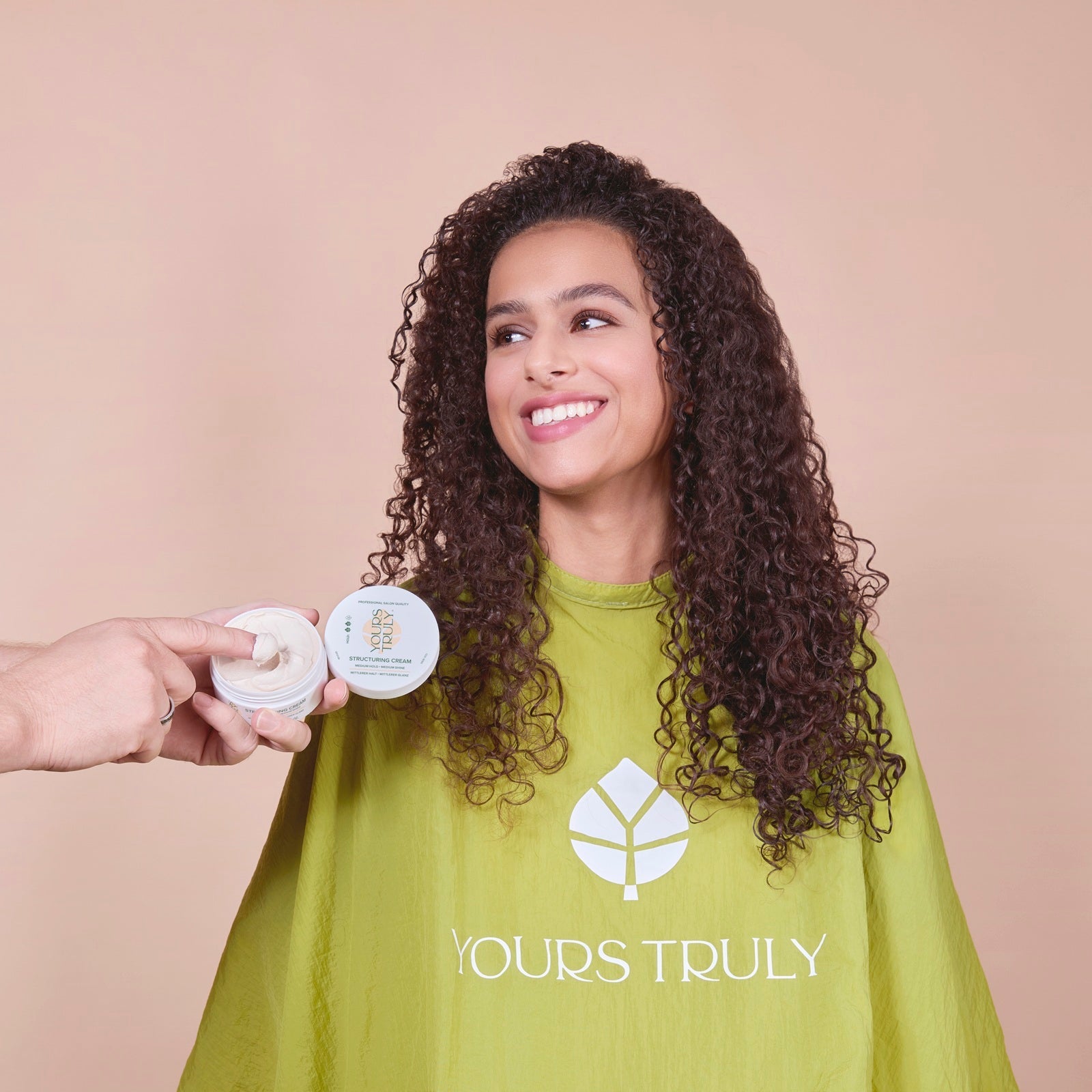 frau-mit-langen-dunklen-lockigen-haaren-trägt-grünen-friseurumhang-mit-yours-truly-logo-und-lächelt-während-eine-hand-einen-tiegel-mit-styling-creme-hält-und-mit-dem-finger-produkt-entnimmt