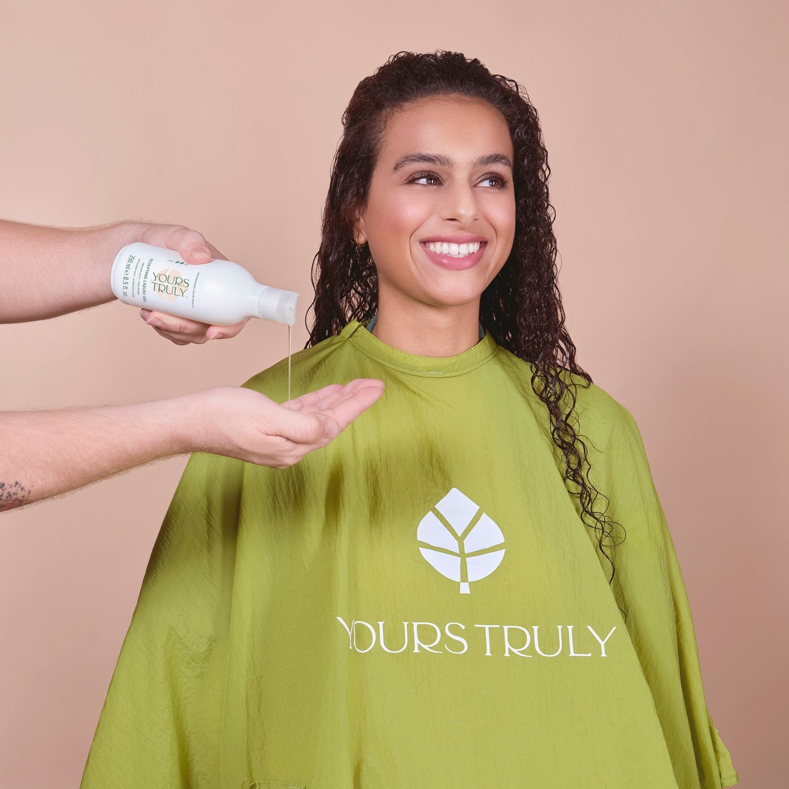 frau-mit-langen-dunklen-locken-trägt-grünen-friseurumhang-mit-yours-truly-logo-und-lächelt-während-eine-hand-gel-aus-weißer-flasche-in-die-handfläche-gibt
