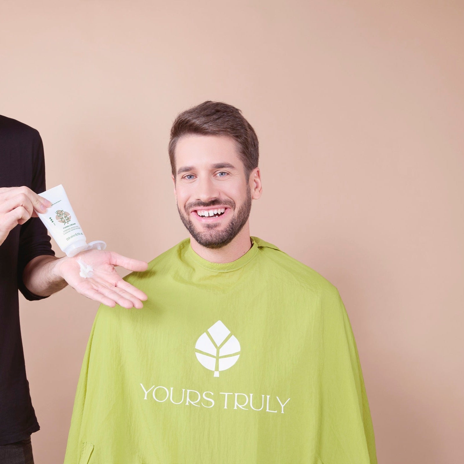 mann-mit-bart-und-kurzhaarfrisur-trägt-grünen-friseurumhang-mit-yours-truly-logo-und-lächelt-während-eine-hand-creme-aus-weißer-tube-in-die-handfläche-drückt