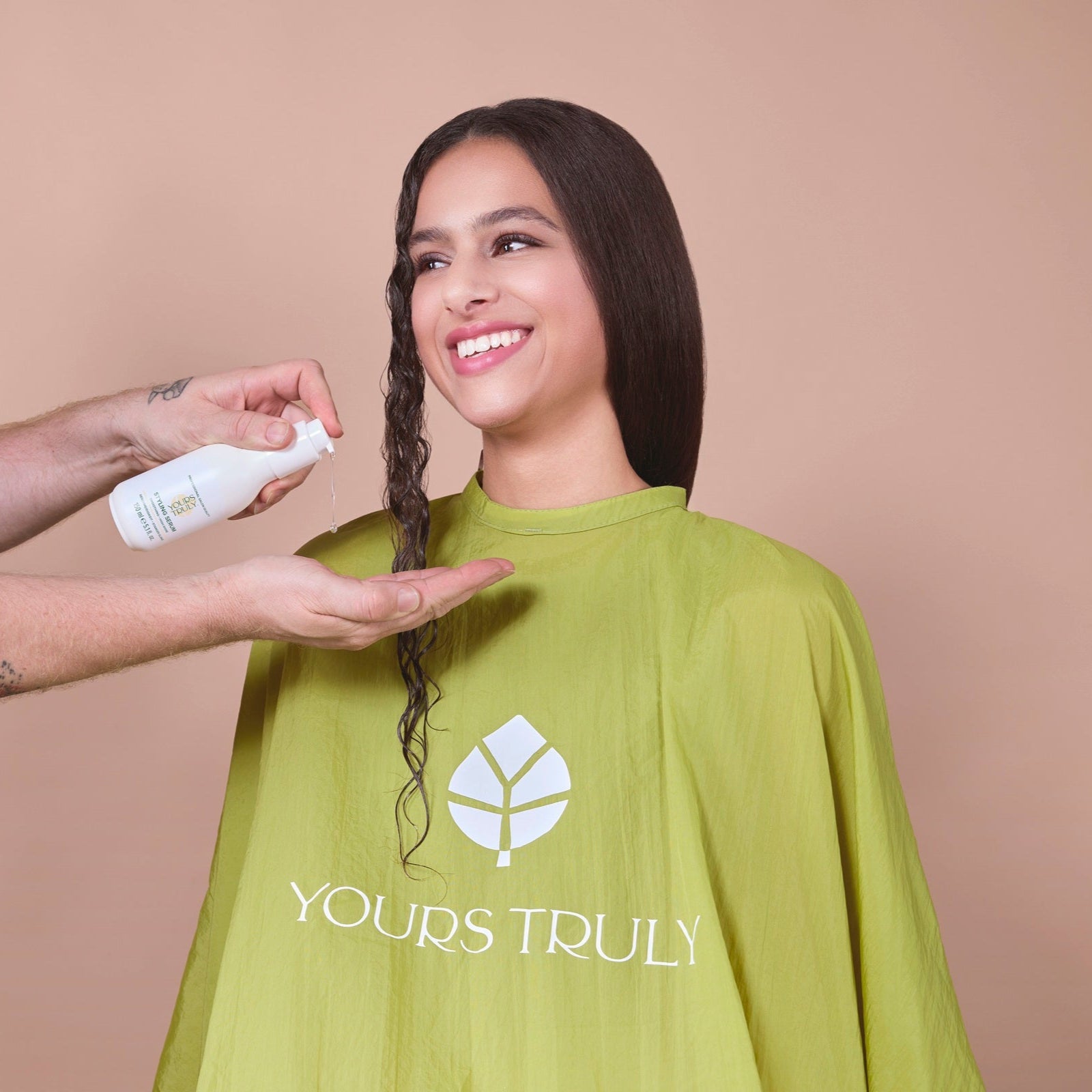 frau-mit-langem-dunklem-haar-trägt-grünen-friseurumhang-mit-yours-truly-logo-und-lächelt-während-eine-hand-produkt-aus-weißer-flasche-in-die-handfläche-und-auf-eine-haarsträhne-gibt