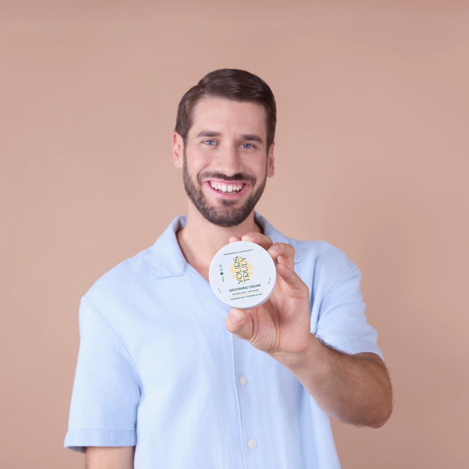 mann-mit-bart-und-kurzhaarfrisur-trägt-hellblaues-hemd-und-hält-lächelnd-eine-runde-dose-yours-truly-grooming-cream-in-die-kamera