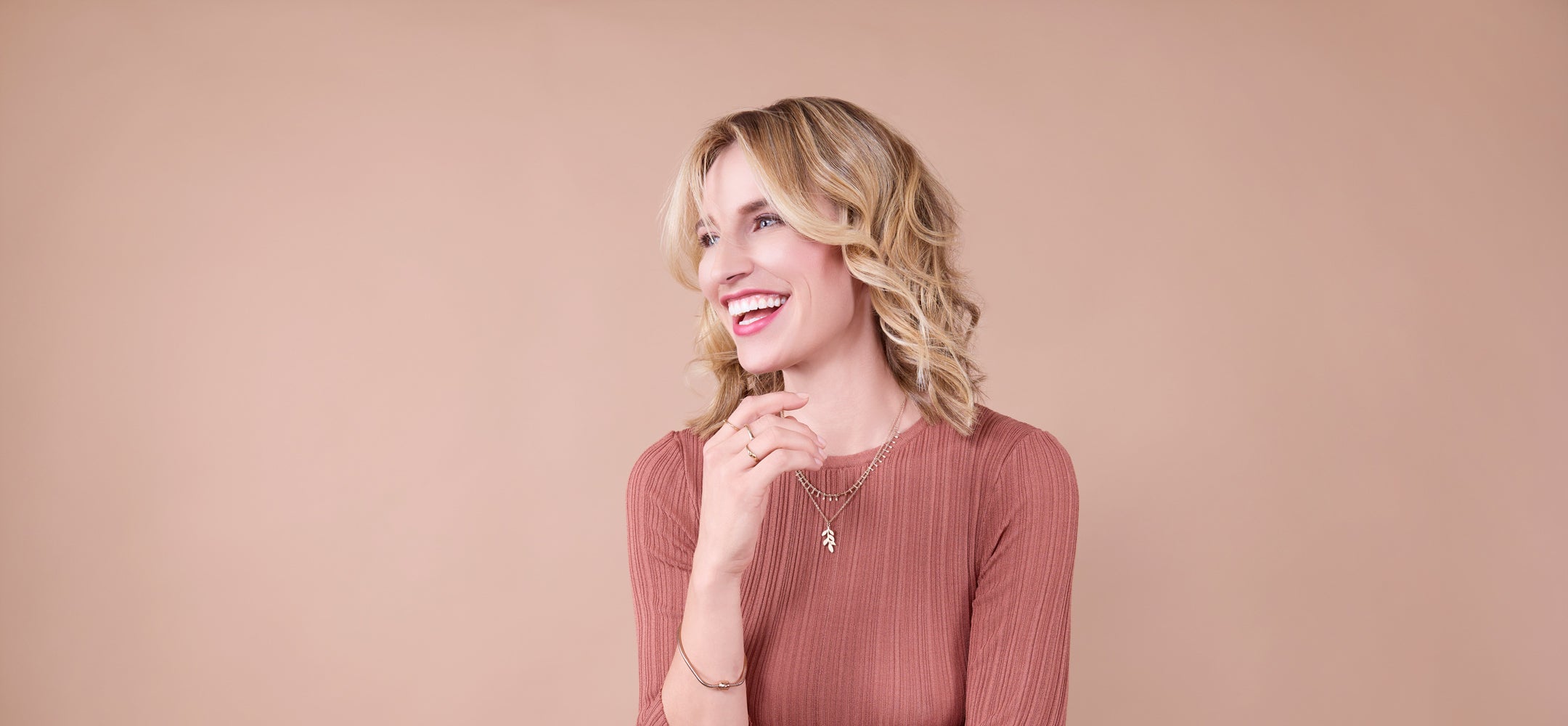 frau-mit-blonden-schulterlangen-locken-und-rostbraunem-oberteil-lacht-und-schaut-nach-links-vor-beigem-hintergrund