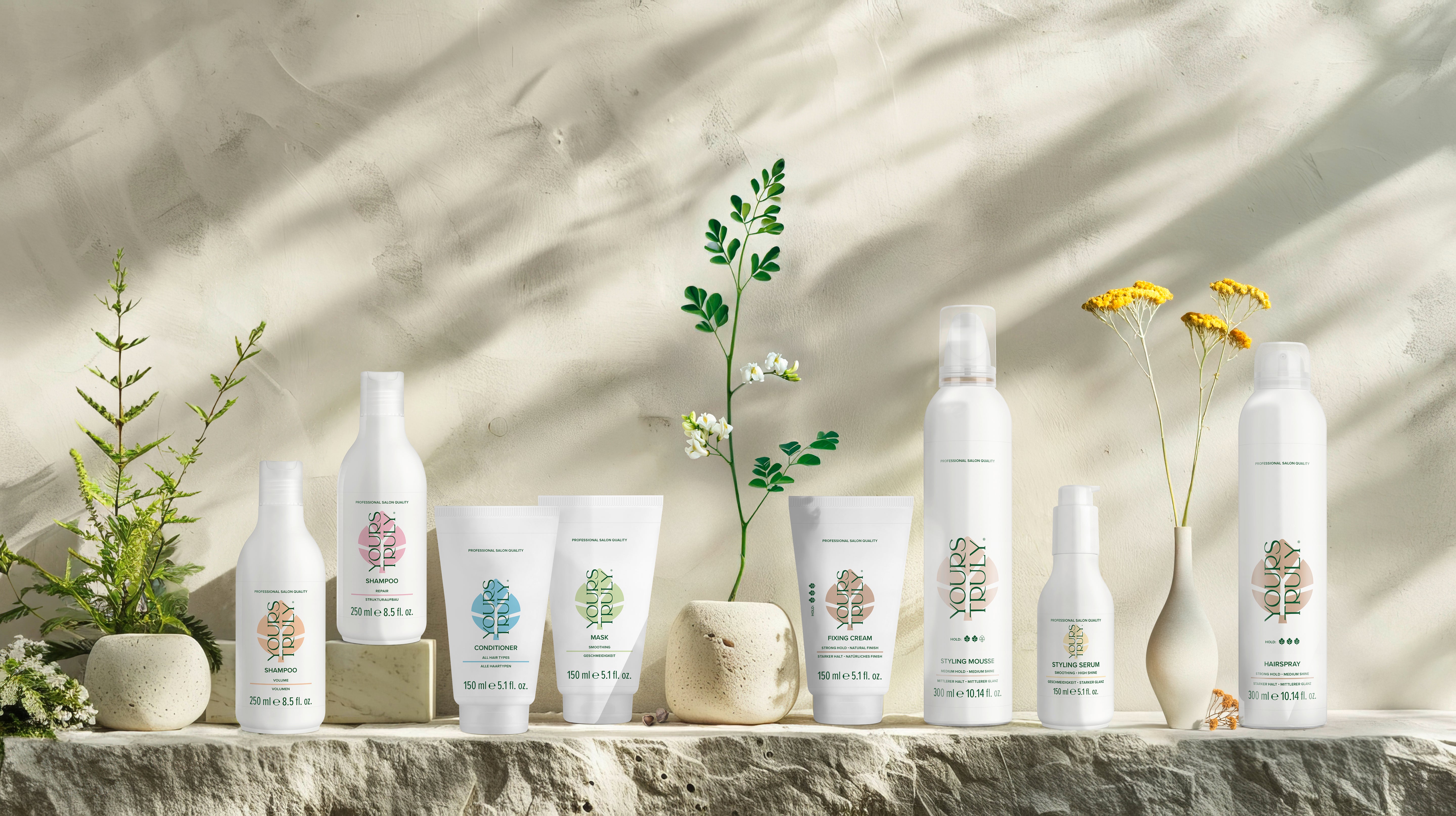 verschiedene-yours-truly-haarpflegeprodukte-wie-shampoo-conditioner-maske-creme-mousse-serum-und-haarspray-auf-naturstein-mit-dekorativen-pflanzen-und-blumen