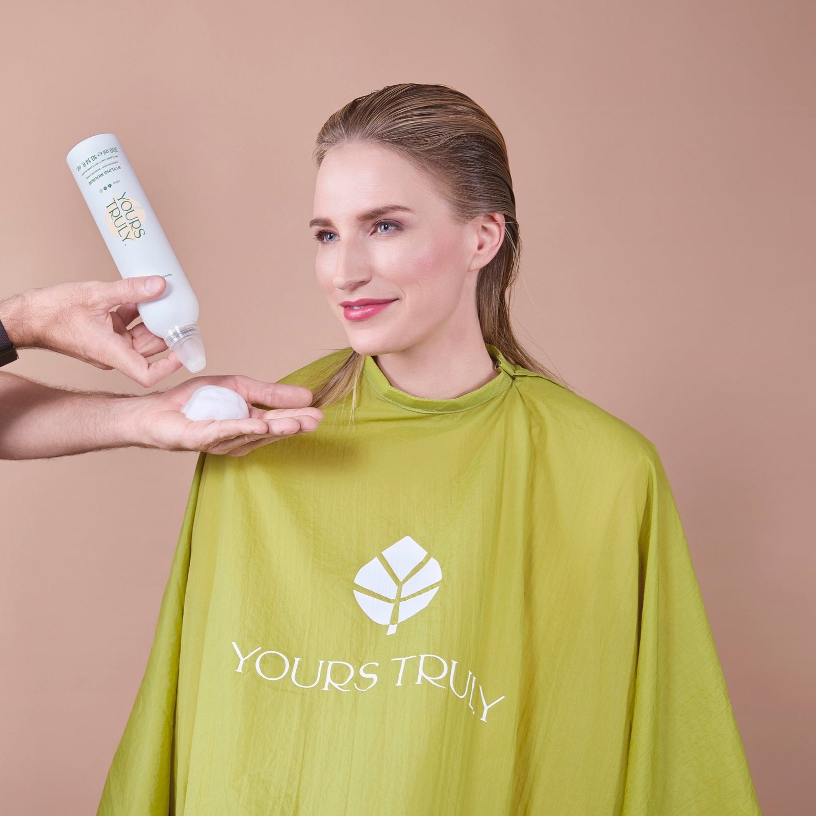 frau-mit-nach-hinten-gekämmtem-blondem-haar-trägt-grünen-friseurumhang-mit-yours-truly-logo-und-lächelt-während-eine-hand-schaumfestiger-aus-weißer-dose-in-die-handfläche-gibt