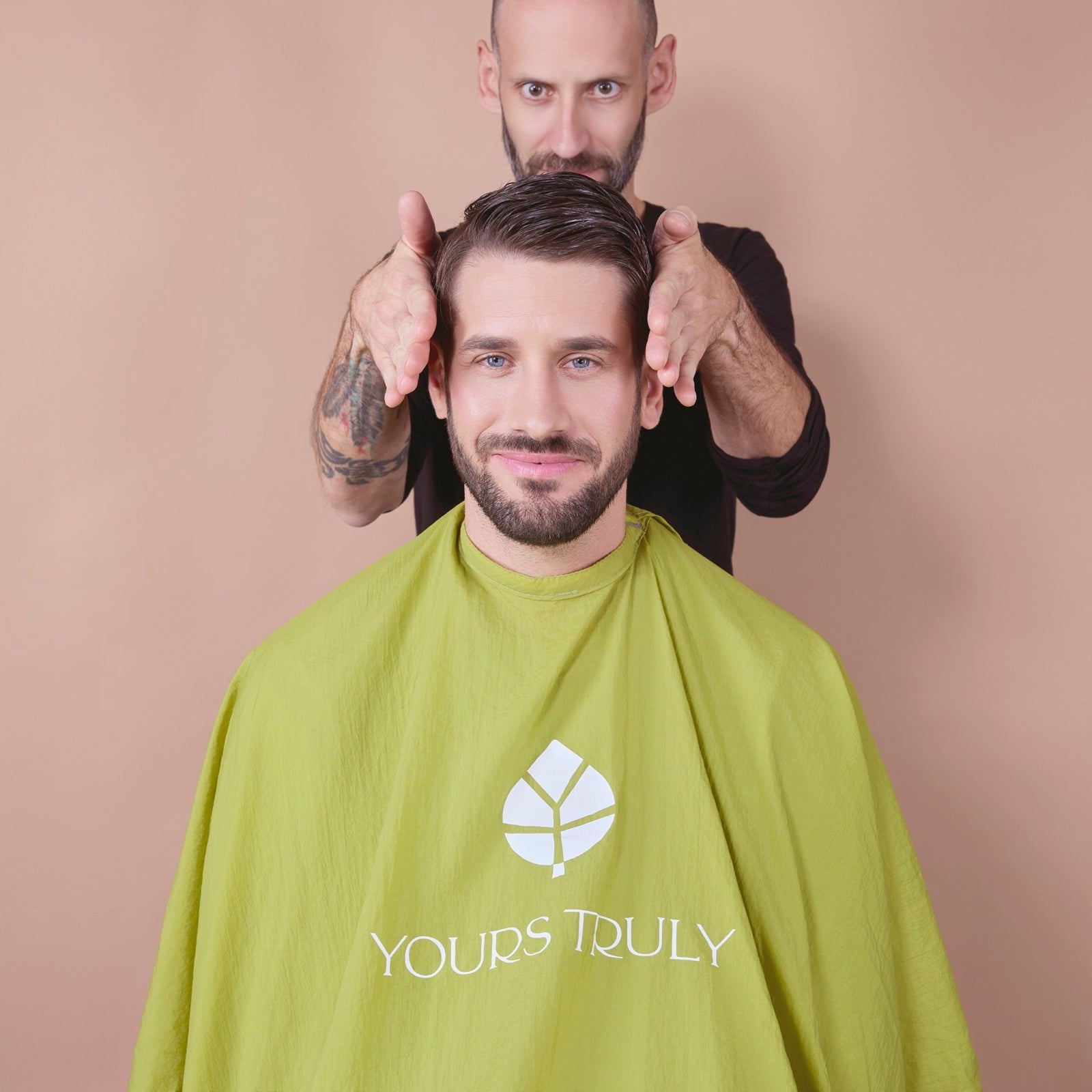 mann-mit-bart-und-kurzhaarfrisur-trägt-grünen-friseurumhang-mit-yours-truly-logo-und-lächelt-während-friseur-mit-beiden-händen-sein-haar-stylt