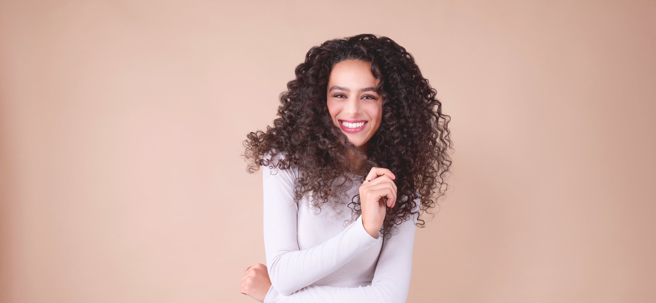 frau-mit-langen-voluminösen-locken-in-weißem-oberteil-lacht-freundlich-vor-beigem-hintergrund