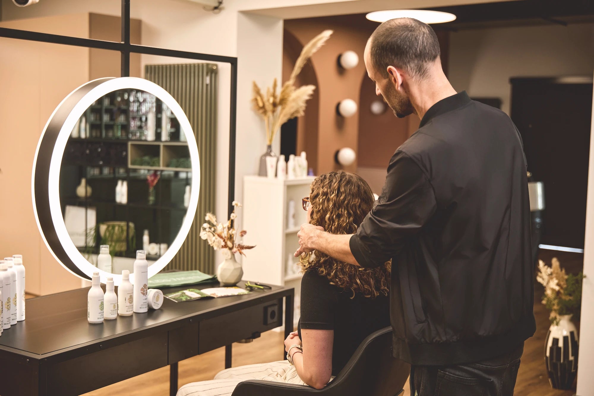 friseur-und-kundin-sitzen-am-frisiertisch-im-salon-und-unterhalten-sich-vor-spiegel-mit-shampooflaschen-und-pflegeprodukten