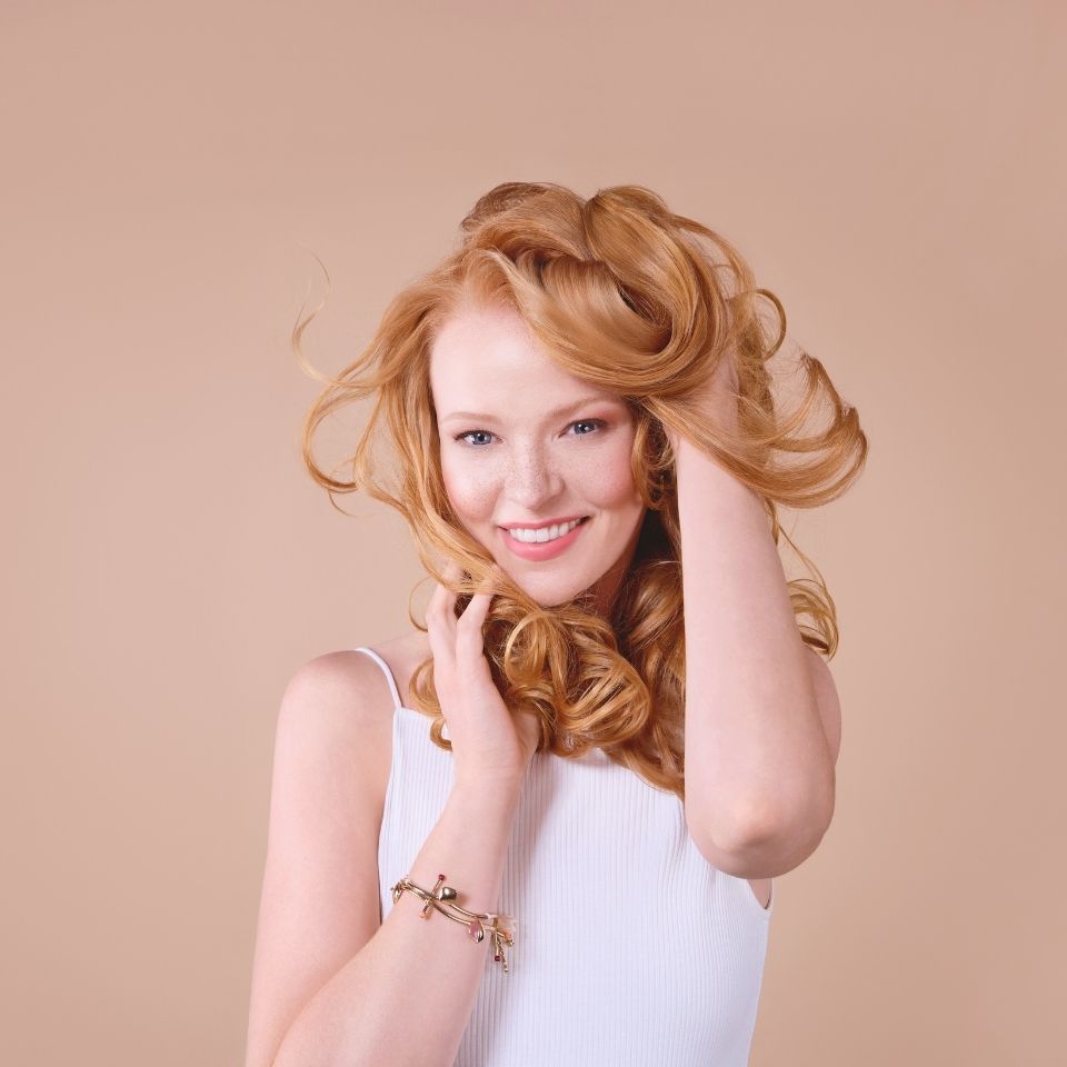 frau-mit-langen-lockigen-roten-haaren-trägt-weißes-oberteil-und-armband-und-lächelt-vor-beigem-hintergrund