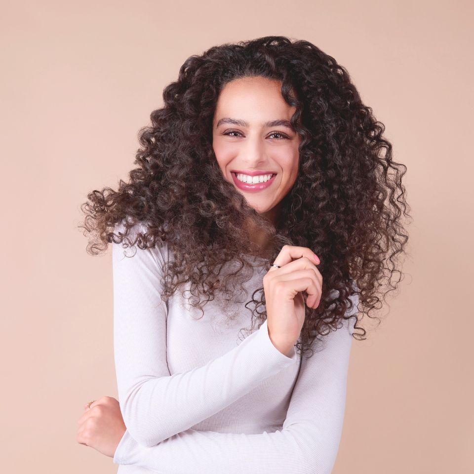 frau-mit-langen-dunklen-locken-trägt-weißes-oberteil-und-lächelt-vor-beigem-hintergrund