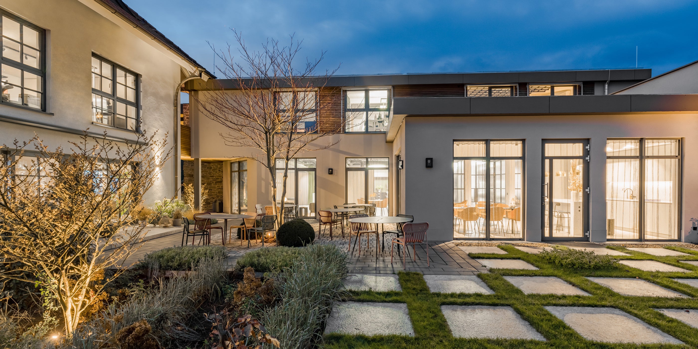 beleuchteter-gebäudeinnenhof-am-abend-mit-großen-fenstern-modernem-garten-quadratischen-steinplatten-und-tischen-mit-stühlen