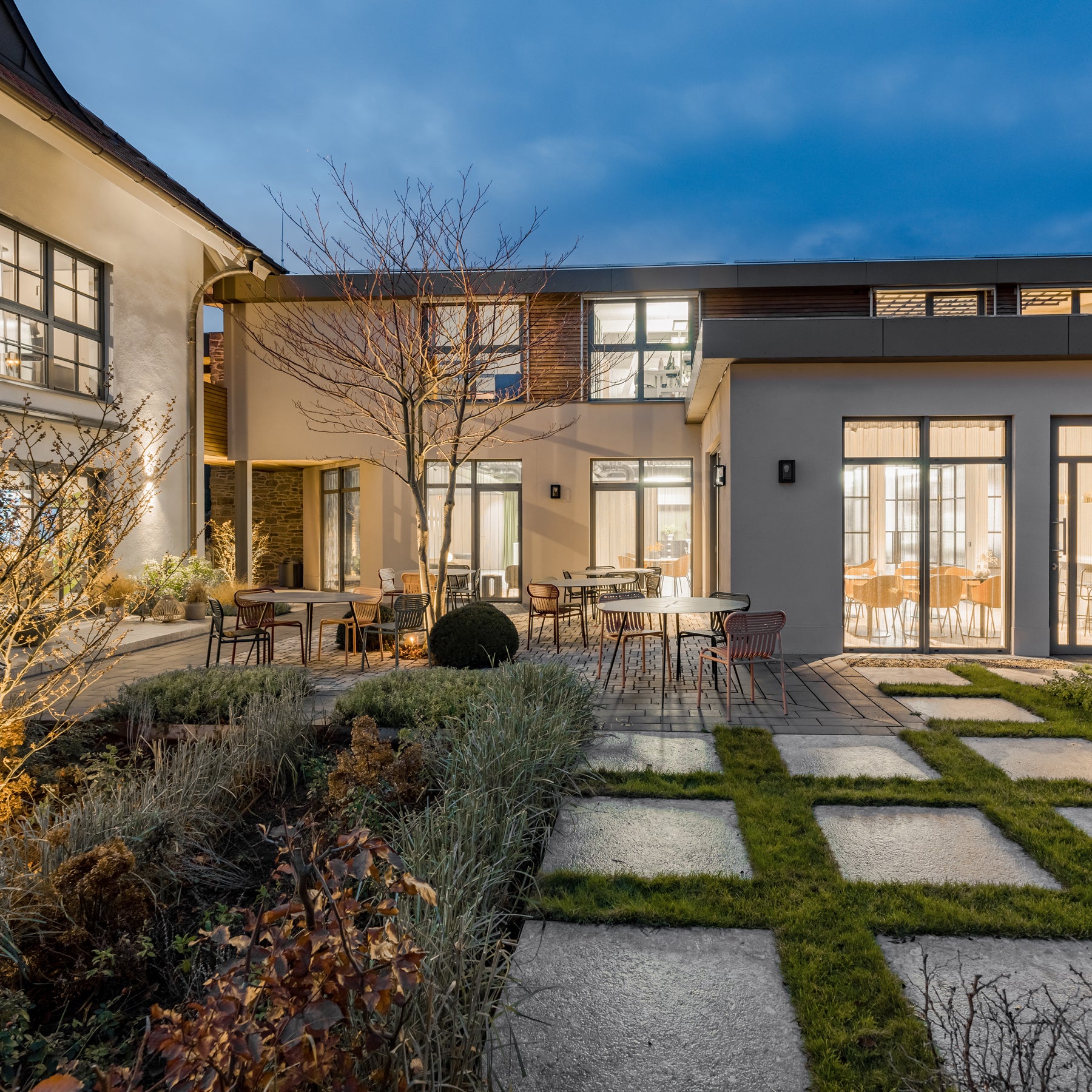 moderner-gebäudeinnenhof-mit-gartenflächen-und-steinwegen-tischen-und-stühlen-im-abendlicht-große-fenster-mit-erleuchtetem-innenbereich